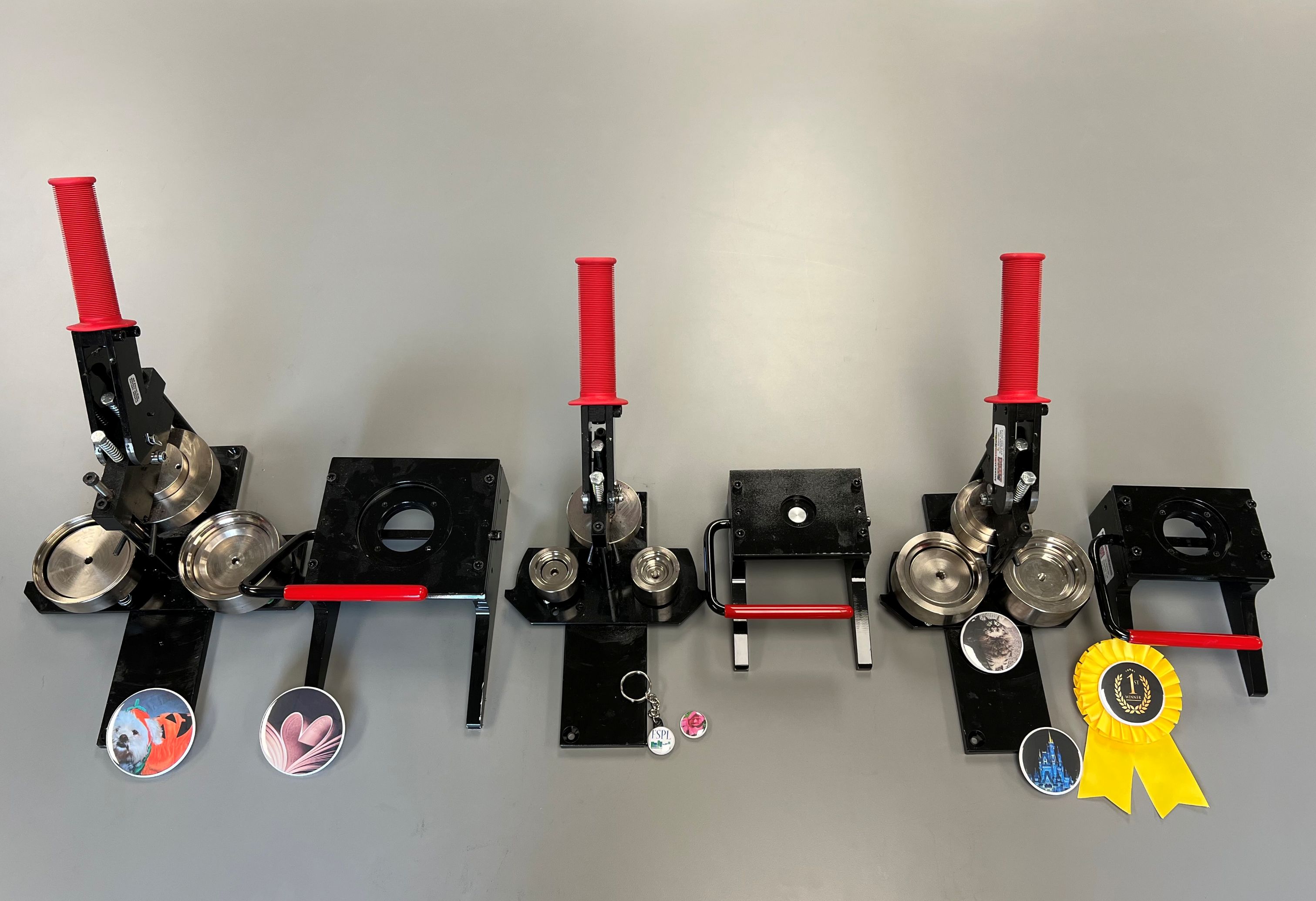 Three button-making machines with red handles on a gray surface, each with circular designs and accessories.