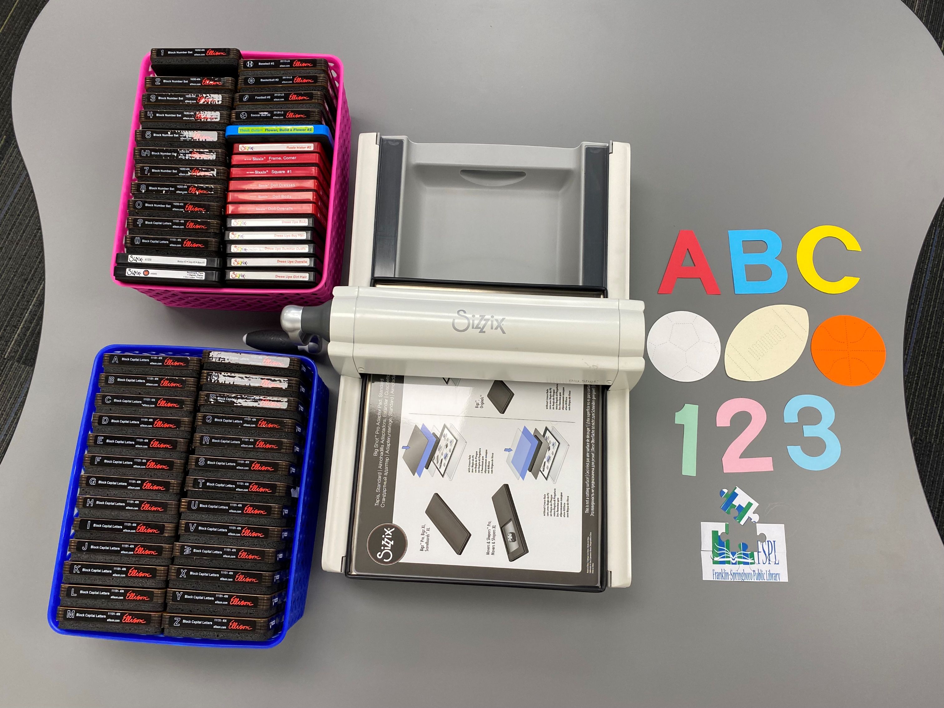 Die-cutting machine with baskets of dies, colorful letters and numbers, and paper cut-outs on a gray table.