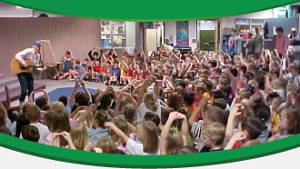A person playing guitar in front of a large group of children sitting on the floor, in a school or library setting.