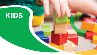 A child's hand stacking colorful wooden blocks with the word "KIDS" in white on a green background.
