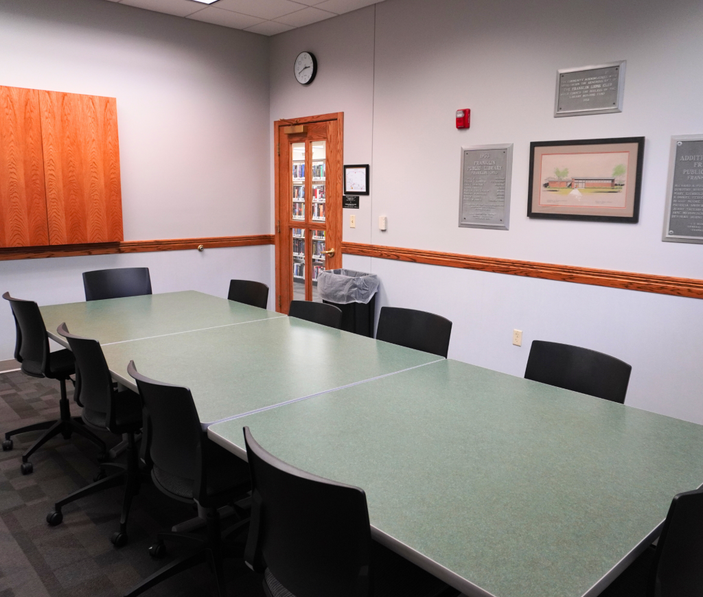 Conference room with a large table, office chairs, wall plaques, a clock, and a wooden cabinet.