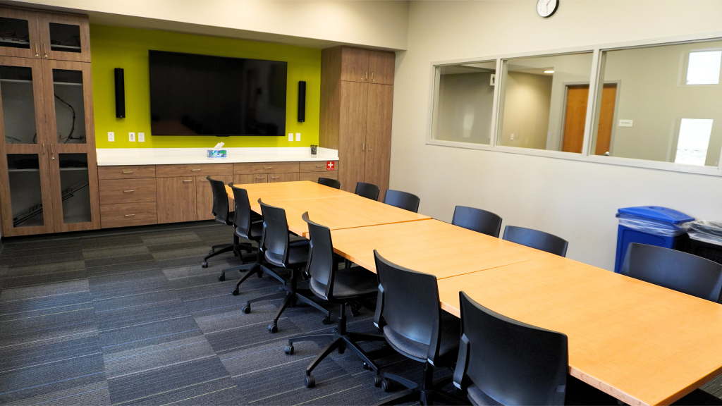 A meeting room with a long table, swivel chairs, a green accent wall, and a large TV.