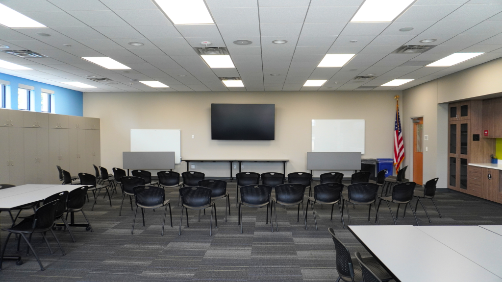 A large meeting room with a TV, whiteboards, black chairs, gray carpet, and light gray cabinets.