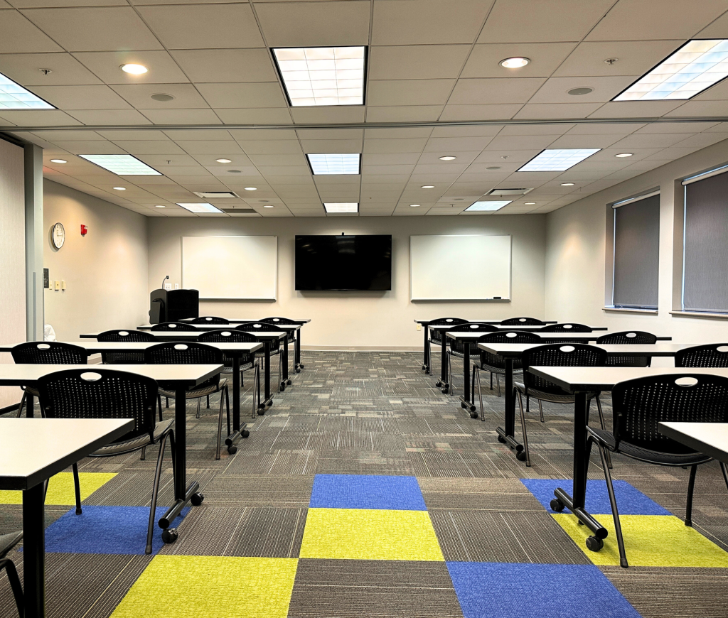 A large meeting room with rows of tables and black chairs, a podium, whiteboards, a large screen, and a checkered carpet of gray, blue, and green squares.