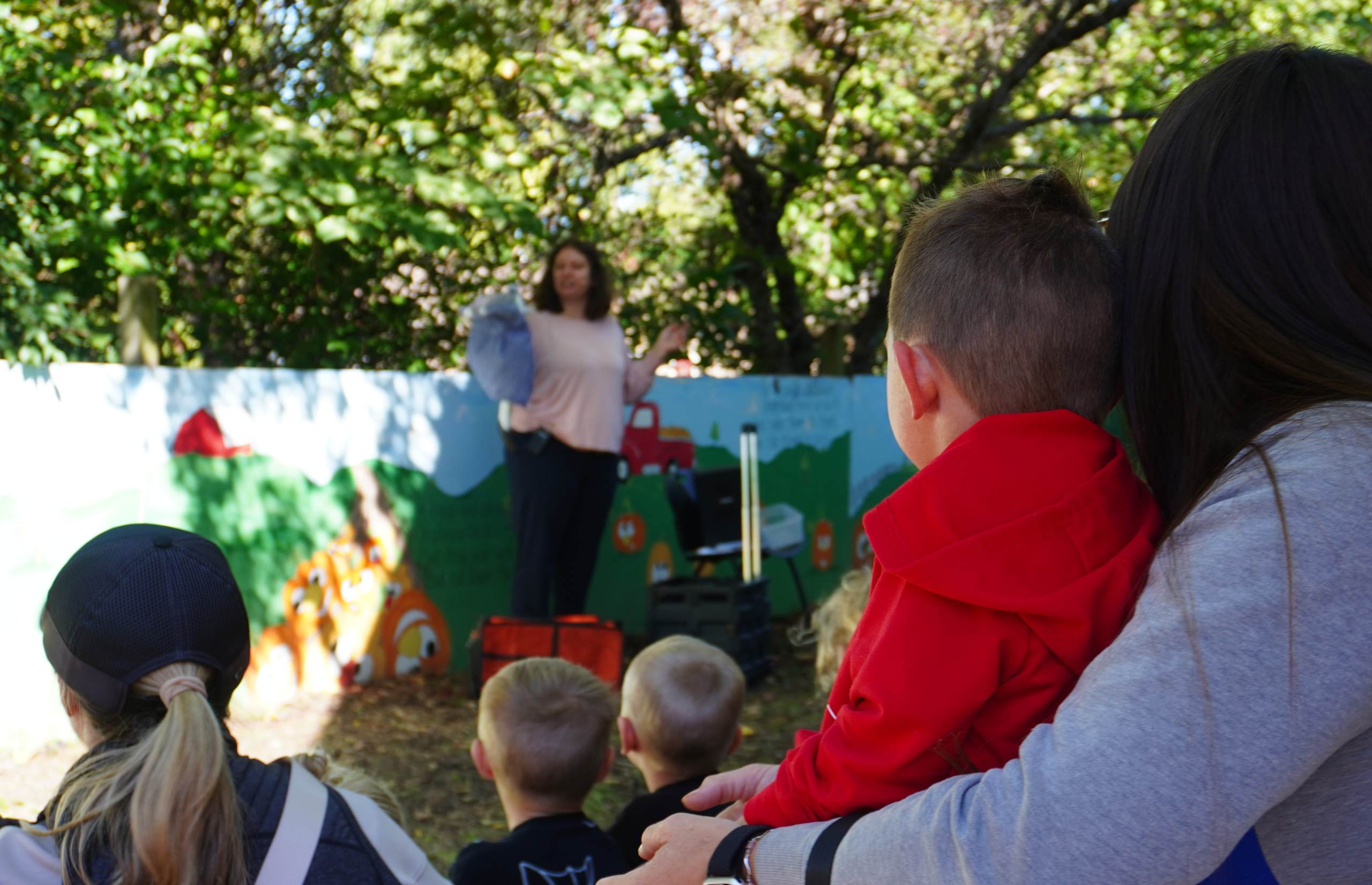 A woman speaks to a group of children outdoors near a colorful mural.