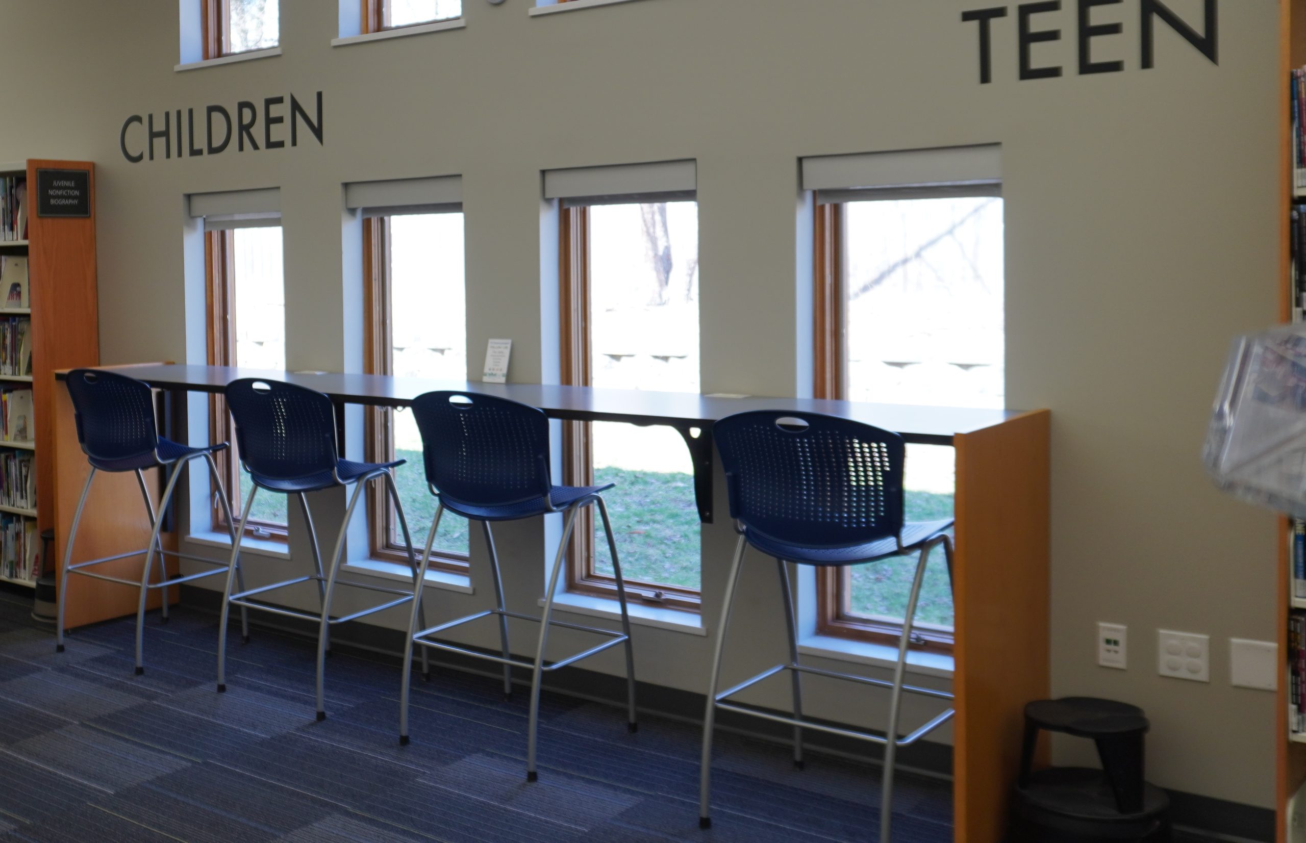 Library area with a high table, blue stools, and large windows, labeled with "CHILDREN" and "TEEN."
